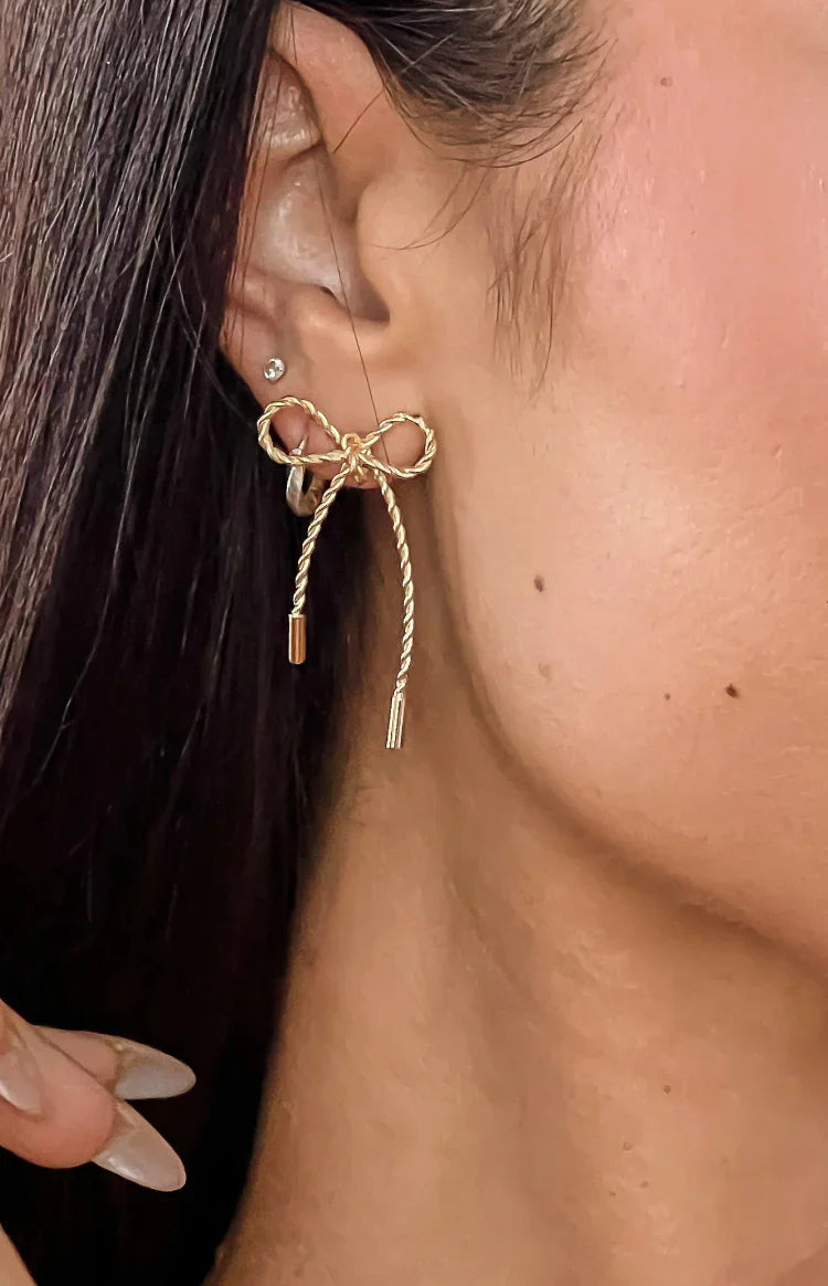 Close-up of a gold twisted metal bow earring with two dangling textured strands, one ending in a cylinder, the other in three